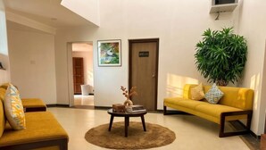 A hotel lobby with a yellow sofa, a round coffee table with decorative items, a large plant, and a door with a sign.
