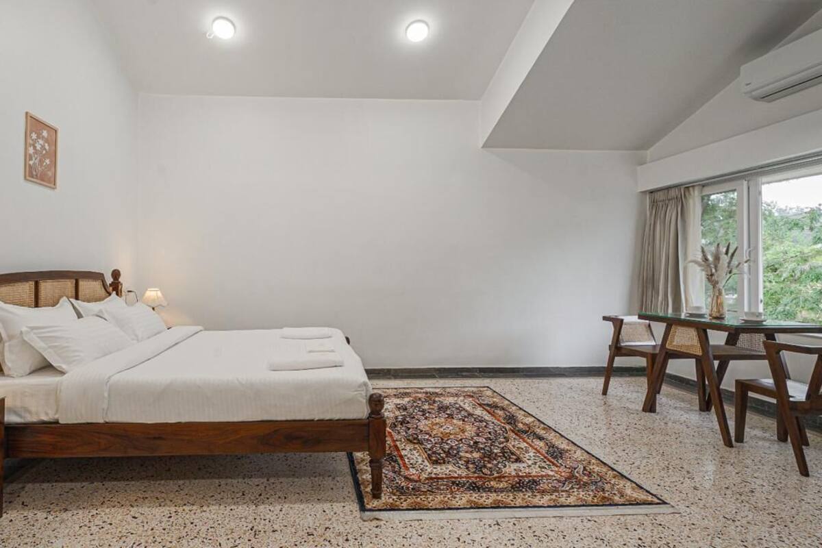 A bedroom with a wooden bed, a bedside table with a lamp, a small table with chairs, and a view of greenery outside.