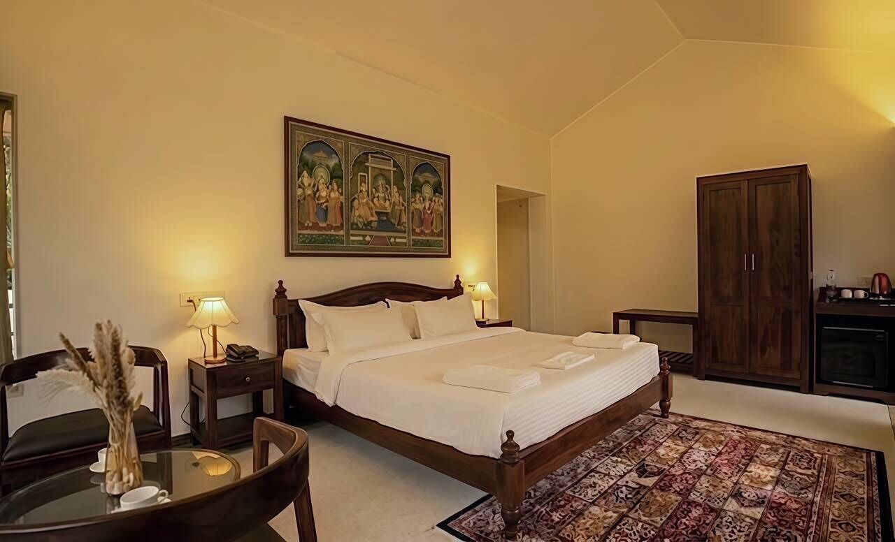 A hotel room with a large bed, bedside lamps, a wooden wardrobe, a desk with a lamp, and a painting on the wall.