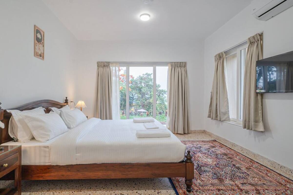 A bedroom with a bed, bedside table, lamp, TV, and a view of the outdoors through a window.