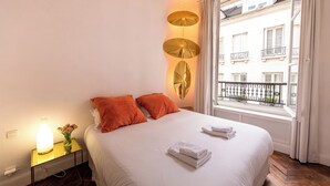 A hotel room with a bed, a bedside table with a lamp, a window with a view of buildings, and a balcony with a railing. - MARAIS, LIVELY, 3BR, REFURB., LIFT, 30–60 days ahead? Private offer FLO-VBp (Paris)