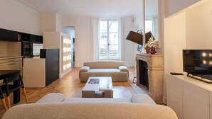 A modern living room with a sofa, coffee table, fireplace, TV, and kitchen area with a fridge and microwave. - MARAIS, LIVELY, 3BR, REFURB., LIFT, 30–60 days ahead? Private offer FLO-VBp (Paris)