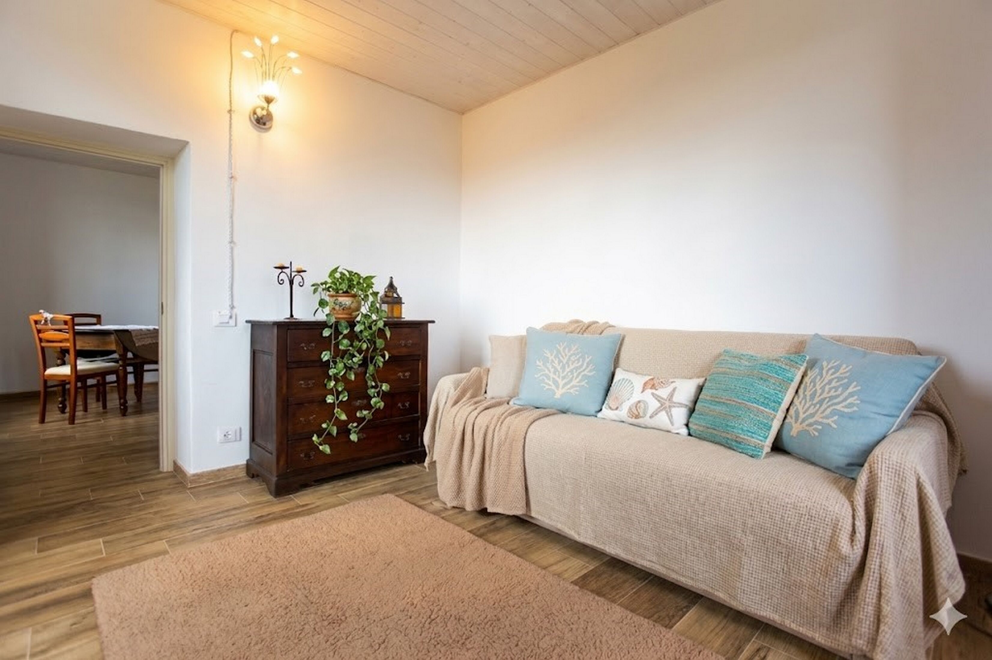Una sala de estar acogedora con piso de madera, un sofá color crema con cojines decorativos, un mueble de madera oscura con una planta y un accesorio de iluminación en la pared.