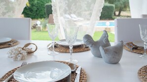 Una mesa dispuesta con platos blancos, cubiertos y copas de vino transparentes, un protector de mesa tejido y una escultura decorativa de un pájaro.