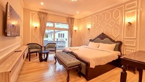 A hotel room with a large bed, two chairs, a small table, a bench, a TV, and a window with curtains. - KING PALACE BY VOYE HOMES (Ooty)