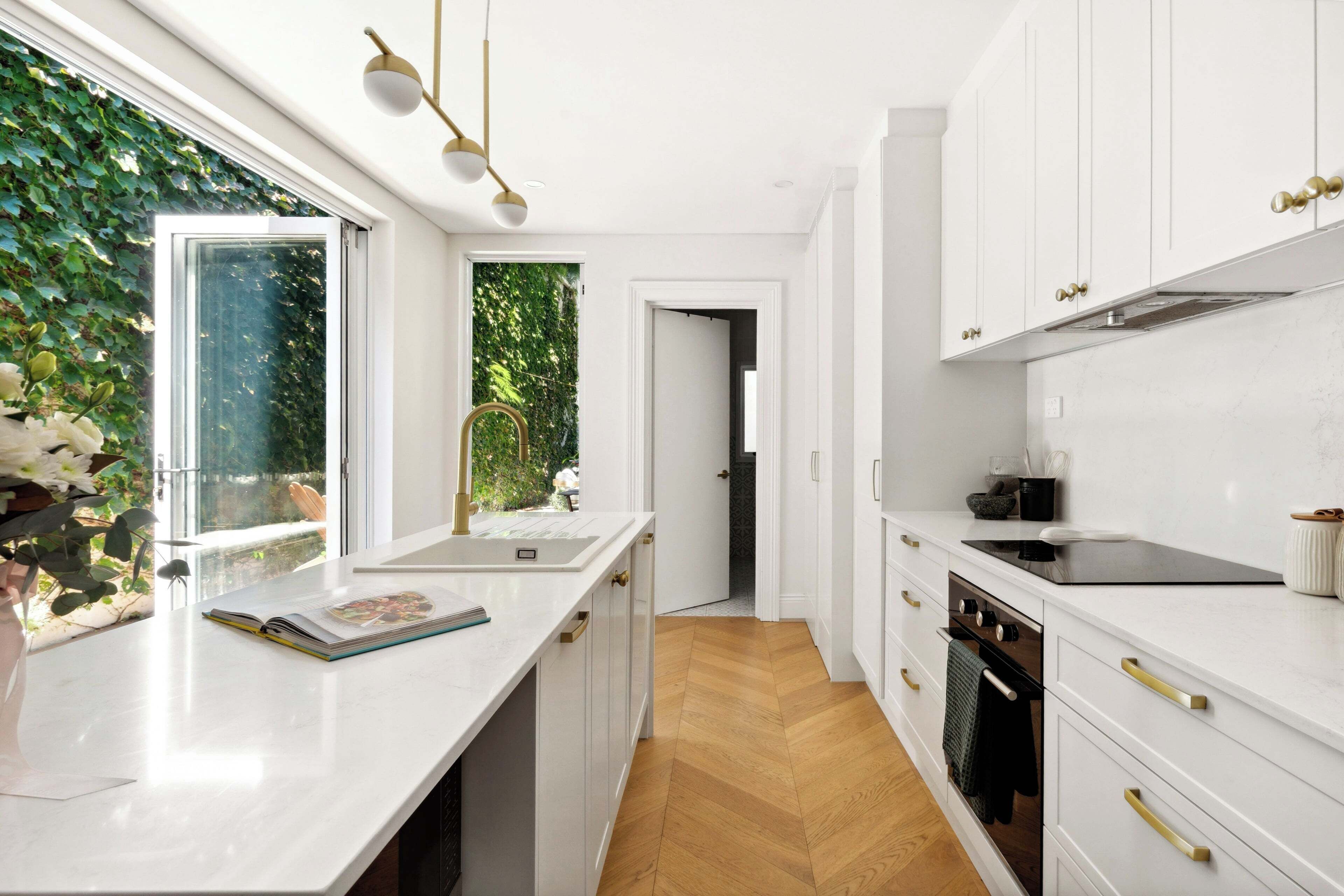 A modern kitchen with white cabinetry, a wooden herringbone floor, and a window with a view of greenery.