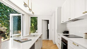 A modern kitchen with white cabinetry, a wooden herringbone floor, and a window with a view of greenery. - Chic Inner-City Oasis in the Heart of Sydney (Redfern)