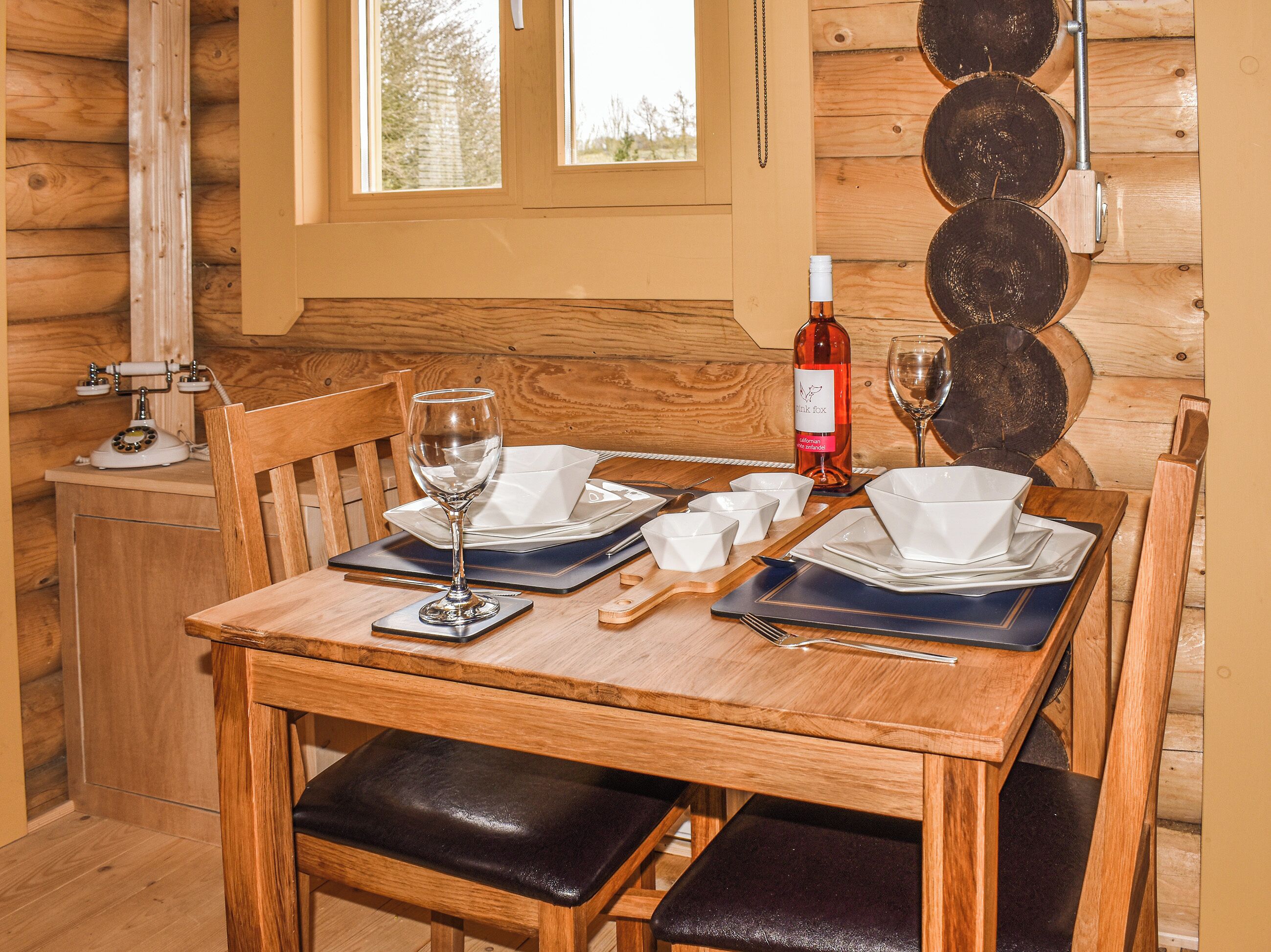 A wooden dining table set with plates, glasses, and a wine bottle, surrounded by wooden chairs and benches.