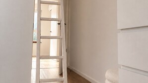 Un couloir avec une échelle blanche, un mur blanc et un meuble blanc orné d’un vase contenant des plantes séchées.