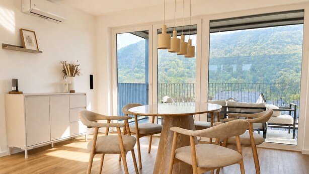 Un espace repas moderne avec une table ronde et des chaises, un meuble blanc et une vue sur un paysage montagneux grâce à de grandes fenêtres.