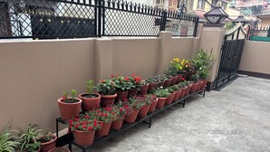 A balcony with potted plants arranged in rows, a concrete floor with hexagonal tiles, and a black metal fence in the background. - Paru home 2bhk Apartment Everest (Kathmandu)
