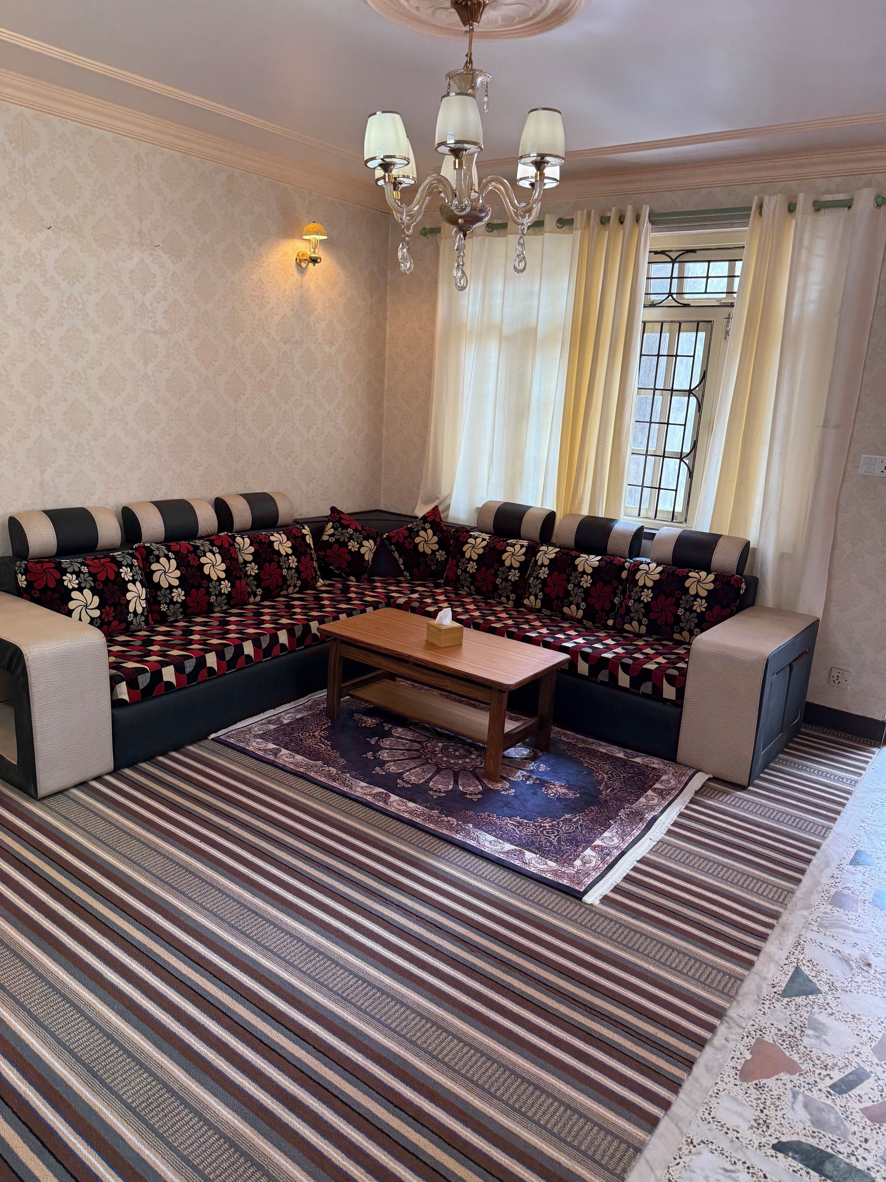 A living room with a sofa, a wooden coffee table, patterned rugs, a chandelier, and a window with curtains.