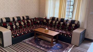A living room with a sofa, a wooden coffee table, patterned rugs, a chandelier, and a window with curtains.