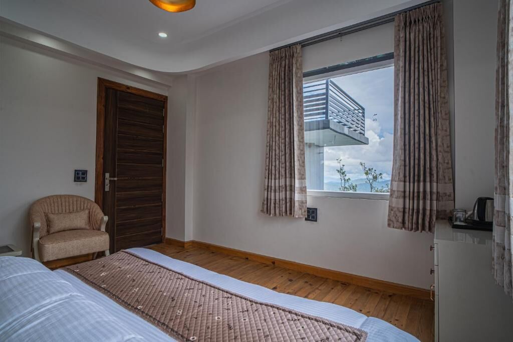 Quarto com cama, uma cadeira, janela com cortinas, piso de madeira e vista para uma varanda.