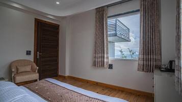 Quarto com cama, uma cadeira, janela com cortinas, piso de madeira e vista para uma varanda.