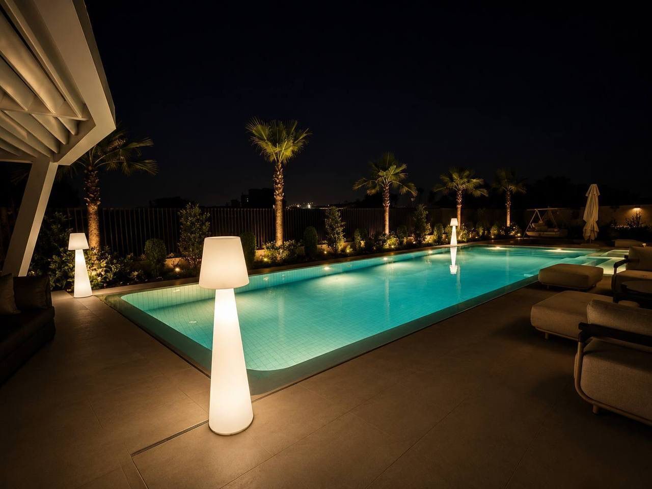 A swimming pool with lounge chairs and lamps, surrounded by palm trees and a fence.