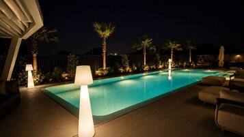 A swimming pool with lounge chairs and lamps, surrounded by palm trees and a fence.