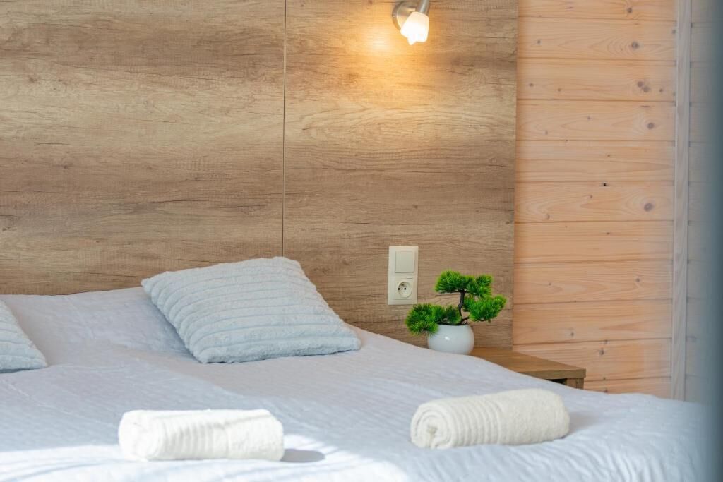 Una cama bien hecha con sábanas blancas y toallas enrolladas, una pequeña planta en maceta sobre una mesita de noche y un aplique de pared.