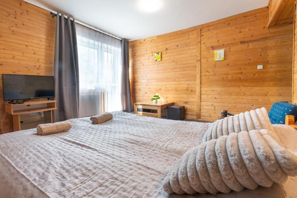 A wooden-paneled room with a bed, a television, a small table, and a window with curtains.