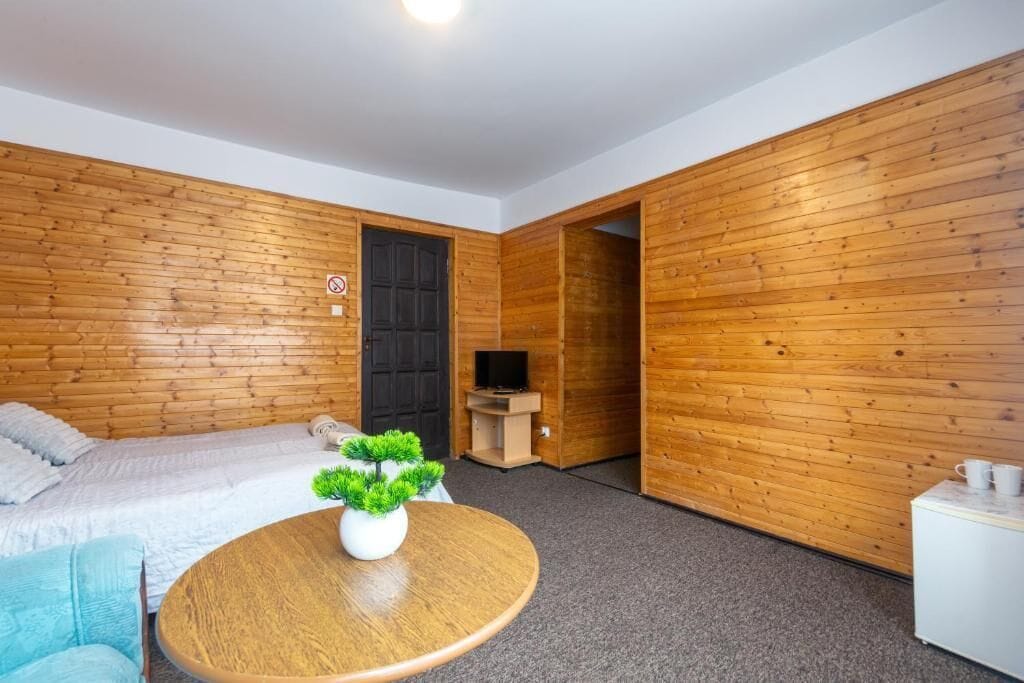 Une pièce aux murs recouverts de lambris en bois, un lit avec une literie blanche, une table ronde en bois ornée d’une plante, et un téléviseur posé sur un meuble.