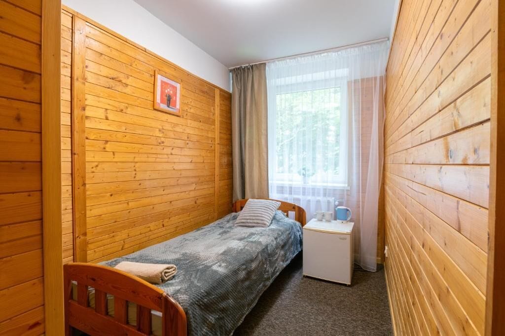 Een kleine eenpersoonskamer met houten lambrisering, een raam met gordijnen en een nachtkastje met een lamp.