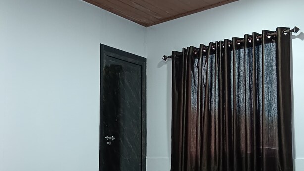 Una habitación con techo de madera, ventilador de techo, puerta negra, ventana con cortinas oscuras, cama con almohada beige y gris, y mesa con camino de mesa estampado.