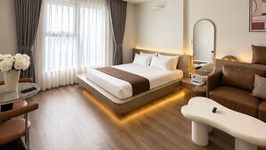 A modern hotel room with a large bed, a sitting area with a sofa and coffee table, a small table with flowers, and a large window with curtains.