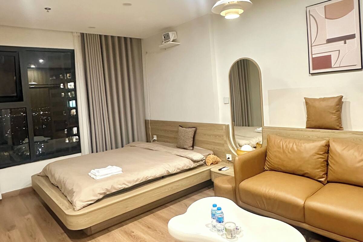 A modern hotel room with a bed, a sofa, a coffee table, a large window, and a city view.