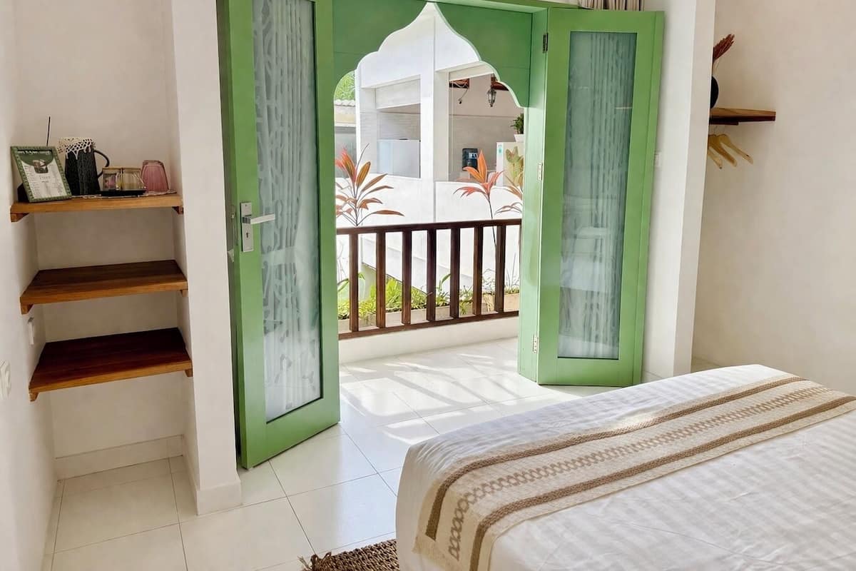 Un dormitorio con una cama, una puerta verde que da a un balcón y un estante con objetos.