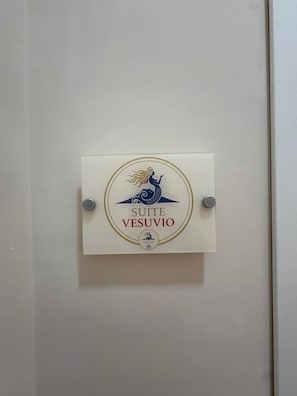 Uma placa quadrada com ilustração de uma sereia e o texto 'SUITE VESUVIO' em uma parede branca.