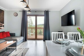 Una sala de estar moderna con un sofá, una mesa de comedor y sillas, un televisor de pantalla plana y un amplio ventanal con cortinas.