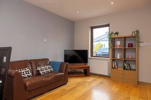 A living room with a brown leather sofa, a flat-screen TV, a wooden bookshelf, and a window with a view of the outside. - Coastal town house beside train and buses only 5 kilometer's from city centre (Blackrock)