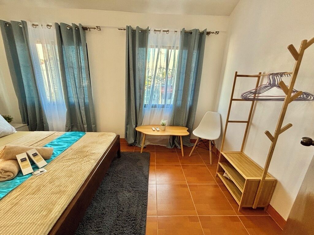 Een hotelkamer met een bed, een tafeltje, een stoel, een kapstok en een raam met gordijnen.
