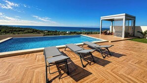 Ein Swimmingpool mit Liegestühlen, einer überdachten Sitzgelegenheit und Blick aufs Meer.