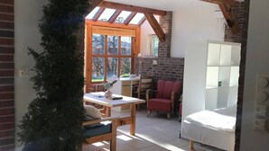 Una habitación con suelo de baldosas, un banco de madera, una mesa con un florero, una planta en maceta, una silla roja, una cama y una pared de ladrillo.