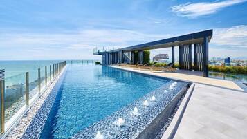 A modern infinity pool with a glass barrier, a covered seating area, and a view of the sea.