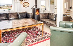 A living room with a large sofa, a glass-top coffee table, a patterned rug, a green armchair, and a wood-burning stove. - Nice home in Thisted with WiFi (Thisted)