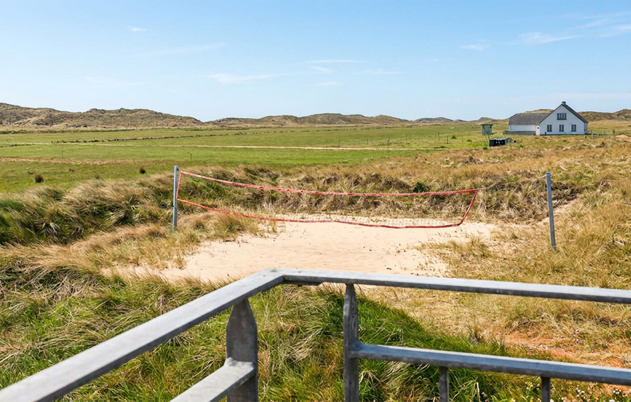 Paisaje rural con una casa blanca, una zona arenosa con una barrera de cuerda y una baranda metálica en primer plano.