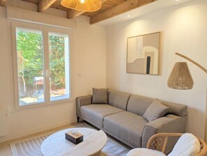 A modern living room with a sofa, a round coffee table, and a wicker chair. There is a large window providing natural light and a view of greenery outside. - Maison "Lomy" bord de mer- 2ch- Large enclosed garden (Pléneuf-Val-André)