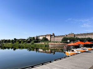 Un fiume tranquillo con barche ormeggiate a un molo di legno, un grande edificio storico sullo sfondo e un cielo azzurro limpido.
