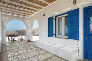 A covered outdoor area with a table and chairs, a window with blue shutters, and a view of the landscape.