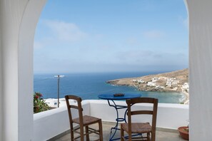 A balcony with two wooden chairs and a small round table, overlooking a coastal landscape with buildings and the sea.