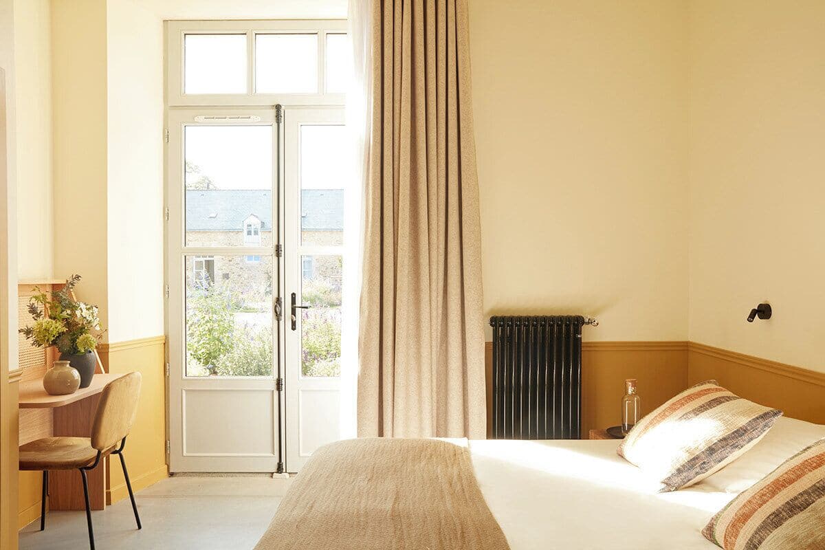 Habitación de hotel con cama, escritorio con silla, radiador y vista por una puerta.