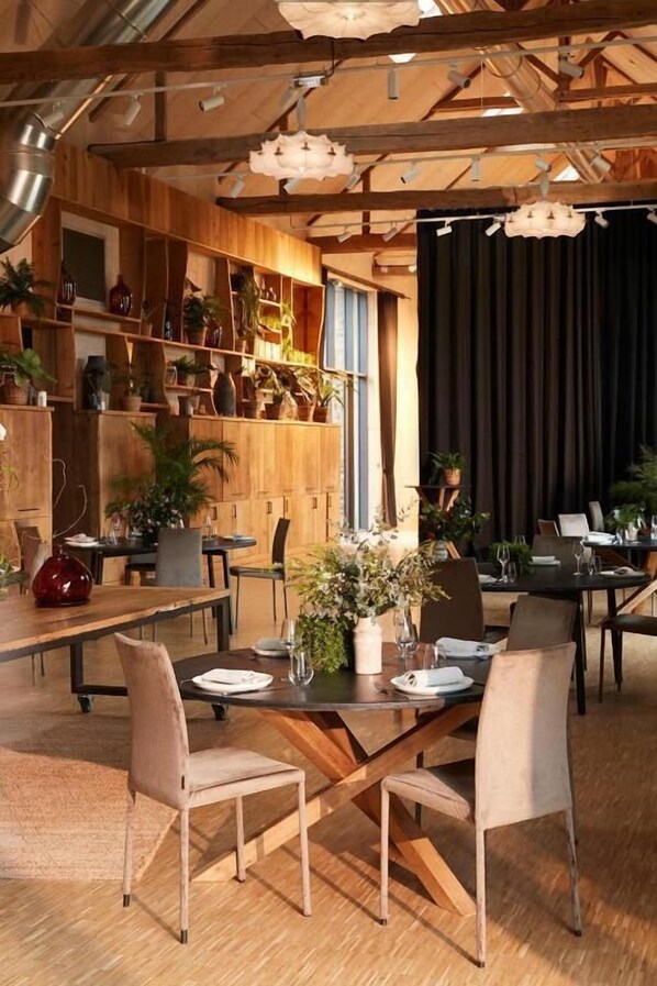 Un espace repas avec des tables et des chaises en bois, un fond constitué d’un rideau noir et des étagères ornées de plantes en pot.