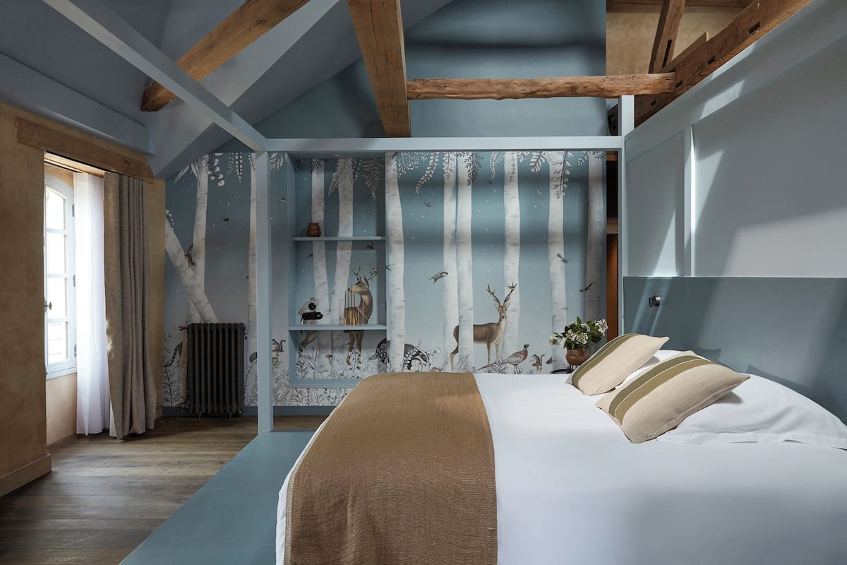 Un dormitorio con cama, ventana con cortinas, radiador y papel tapiz con motivos naturales.