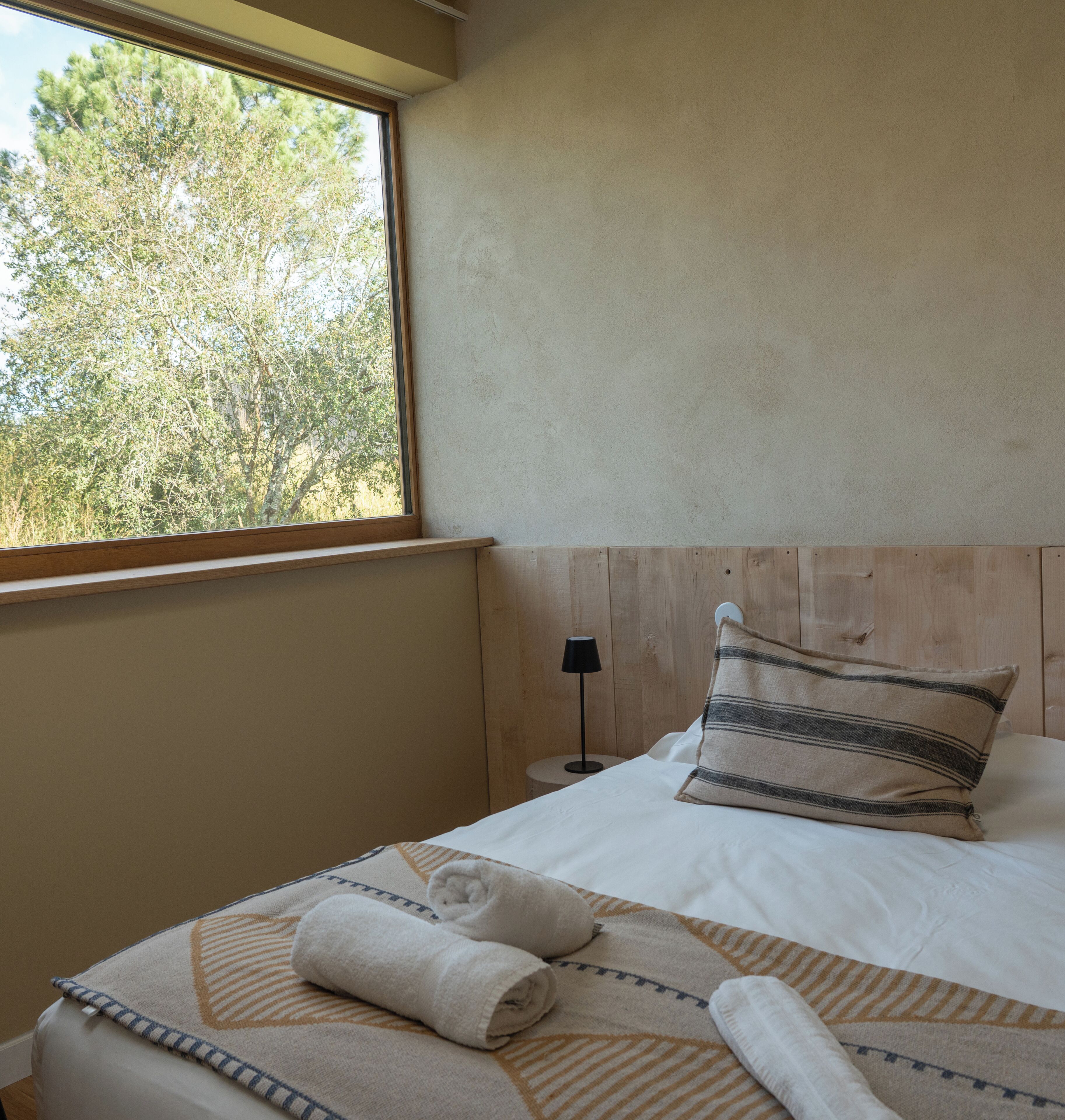 Ein Schlafzimmer mit großem Fenster, einem Bett mit gestreiftem Kopfkissen und zwei aufgerollten Handtüchern auf dem Bett.