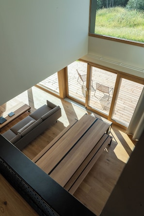 Quarto moderno com piso de madeira, um sofá, uma mesa e uma janela grande com vista para um deck de madeira.