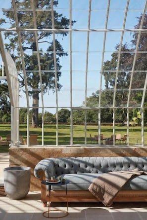 Ein Wintergarten mit gepolsterten Sitzgelegenheiten, einem großen Fenster und Blick auf eine Rasenfläche.