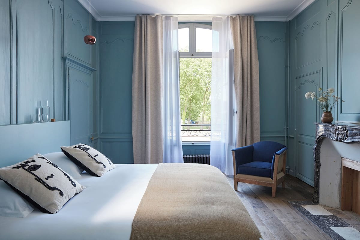 Un dormitorio con cama, una silla, una ventana con cortinas y una chimenea.