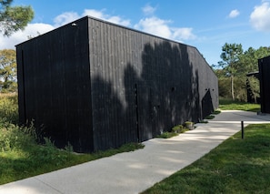 Un grand bâtiment noir, doté d’un toit plat, avec une allée pavée menant à l’entrée et une zone herbeuse bordée d’arbres en arrière-plan.